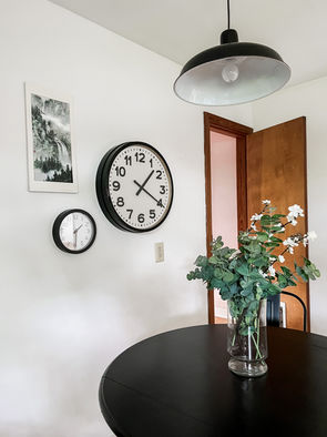A modern farmhouse style black metal pendant light hangs over a round black table and chairs.  Two clocks on the wall look so nice with the black and white painting.
