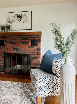 Brick fireplace, white and black polka dot side chair, oversize white pot with greenery, grey and white floral wall art