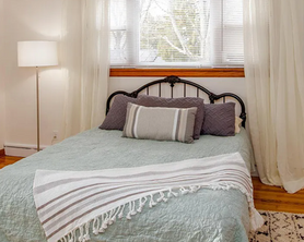 Bedroom staged by Souled Home Design in State College, PA. Black metal headboard, pastel blue-green coverlet on bed with grey and cream striped blanket draped at the bottom of the bed. Grey and white striped pillow on the bed, sheer curtains.