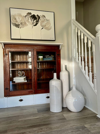 Port Matilda Home staged by Souled Home Design. Photo features a wooden built-in shelving unit with glass doors that is staged with books. Large white pots are on the floor in front of the shelves, the floor is grey washed Luxury Vinyl, a grey and white floral print is hanging over the shelves