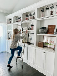 White built in shelving unit with library ladder.  Designer, Heather Haas of Souled Home Design is demonstrating the custom made library ladder from Etsy.  Shelves decorated with decor items from Pottery Barn.  Glass jars with black decorative tops from Pottery Barn, Decorative books from Pottery Barn.