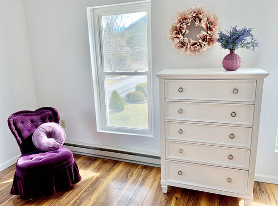 A purple velvet chair with a pink pillow on it look so pretty with the tall white dresser with the pink vase with lavender flowers on it and a pretty floral wreath hanging on the wall behind it.