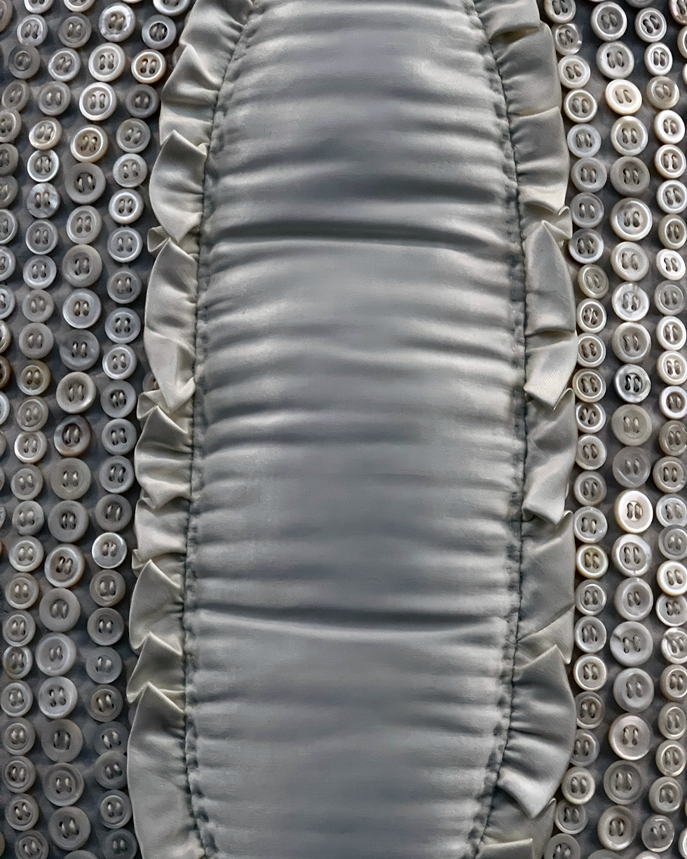 Close-up of a fabric with ruffled edges and white buttons in a neat pattern. The fabric is gray and shiny, giving a textured look.