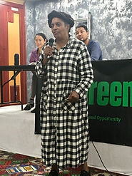 Esther Little Dove, African-American woman in a long black and white checked dress, holds a microphone and speaks at Don't Displace the South End meeting space.