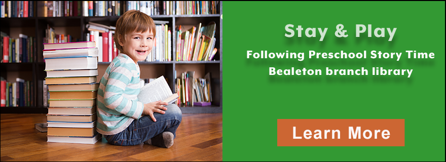 young boy holding an open book in front of bookshelves. Stay and Play following Preschool Story Time, Bealeton branch library