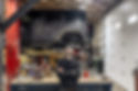 A man smiling with his arms crossed while leaning back against an auto repair workbench with a vehicle and shop in the background