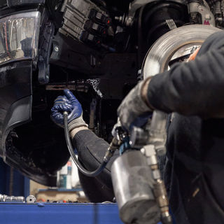 A man reaches up into the wheel well of a vehicle with the tire removed and sprays rust preventing oil