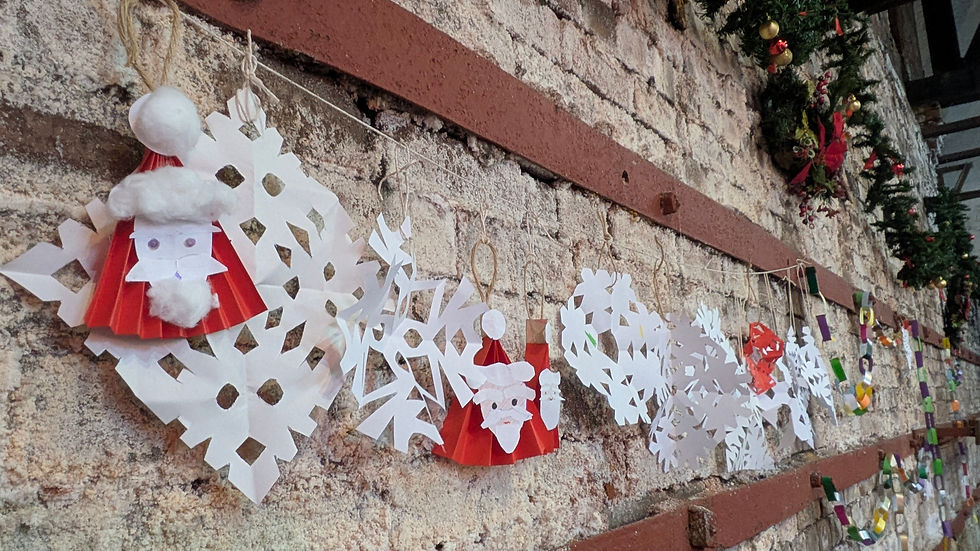 Brick wall with paper decorations hanging from it on string - there are multiple snowflakes - and three santa paper decorations - red paper folded like a fan pinched into a triangle and his face on it - as well as multi coloured paper chains.