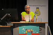 KSLF - Seni Seneviratne close up, stood on stage behind a podium and reading from a piece of paper.