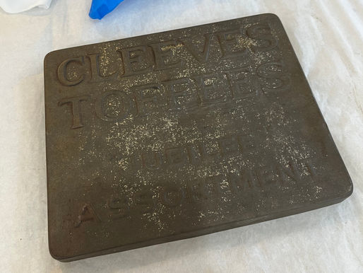 A dusted and waxed toffees tin lid - dark metal with embossed letters that read_ Cleeves Toffees - Jubilee Assortment.