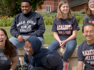 A group of Macalester College students sitting outdoors on campus, smiling and laughing together, all wearing Macalester College shirts.