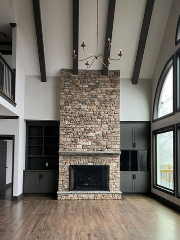 Living room with floor-to-ceiling stone fireplace, vaulted ceiling with dark wood beams, large arched windows, built-in cabinetry, and rich hardwood flooring.