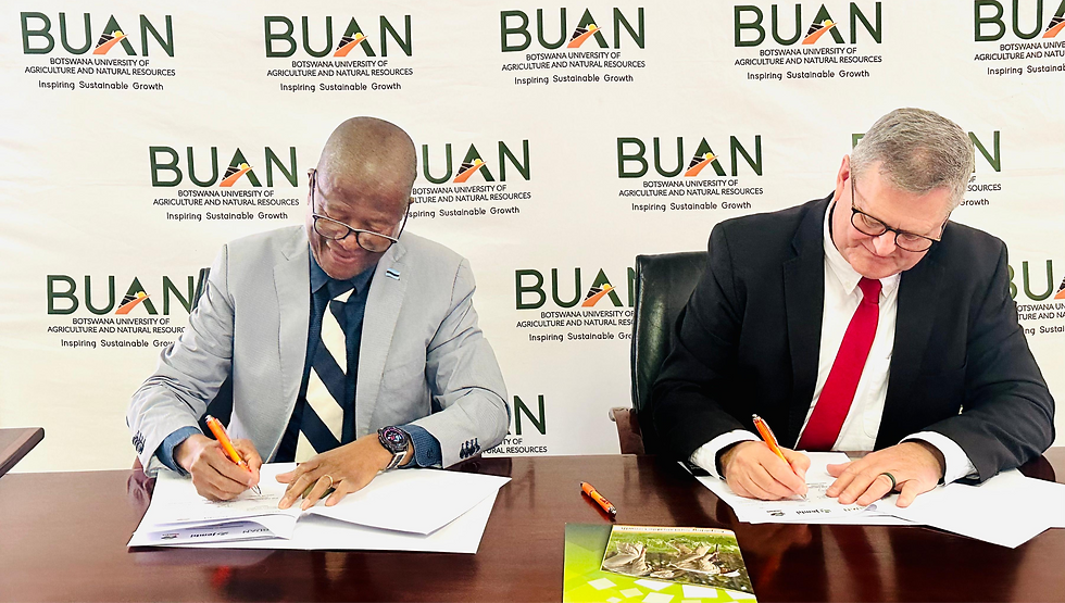 Two men in suits sign documents at a table. Background shows BUAN logo; one wears a gray jacket and striped tie, the other a red tie.