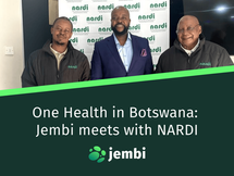 Three men smiling in front of a white "nardi" backdrop. Two wear black jackets; one is in a blue suit. Indoor setting with a tile floor. Title says One Health in Botswana: Jembi meets with NARDI