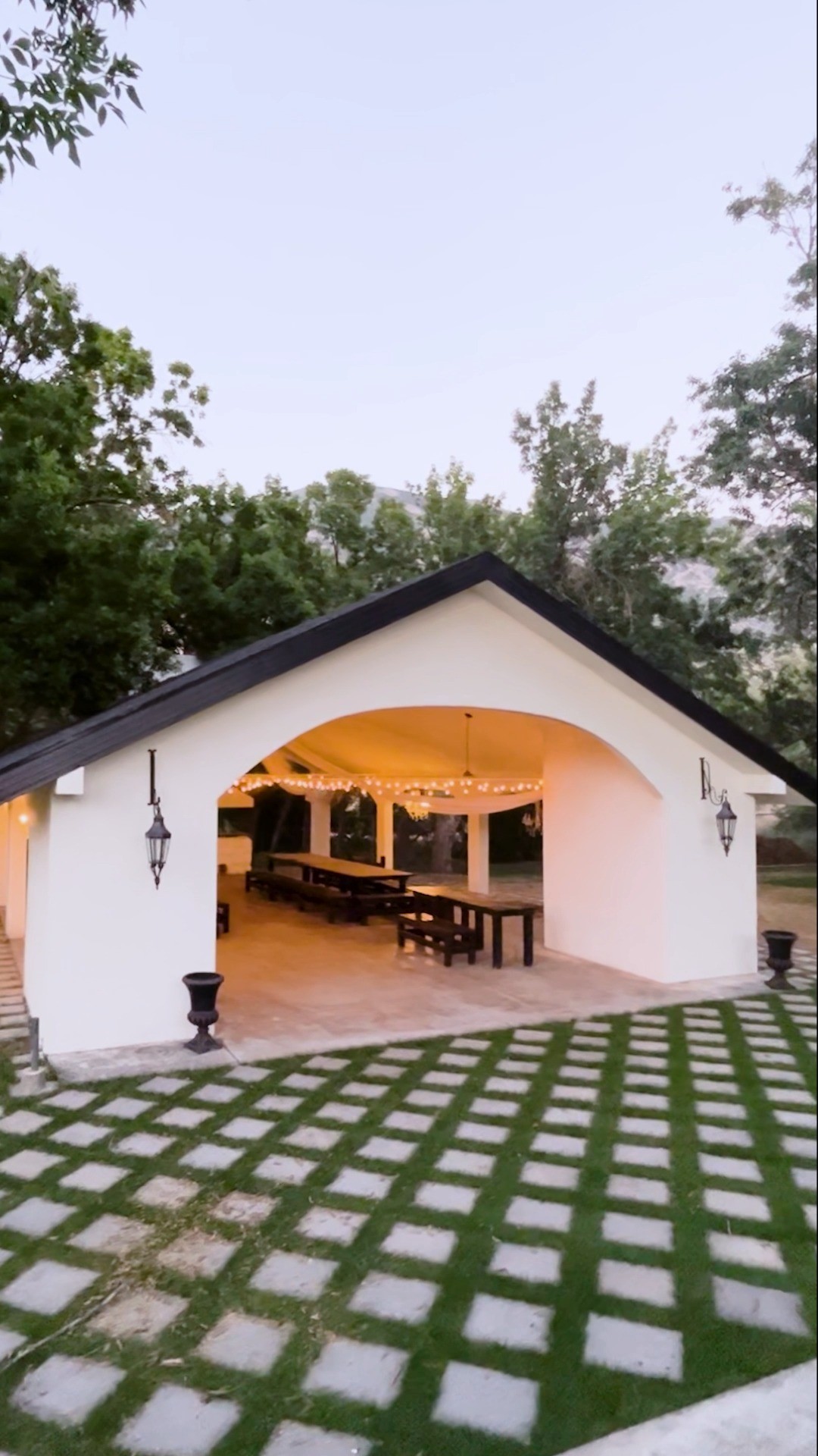 The Hidden Grove at Stone Gate Weddings & Events in Pleasant Grove, Utah: a white stucco open-air pavilion with a dark peaked roof, string lights, and long wooden tables, framed by mature trees and mountains at dusk