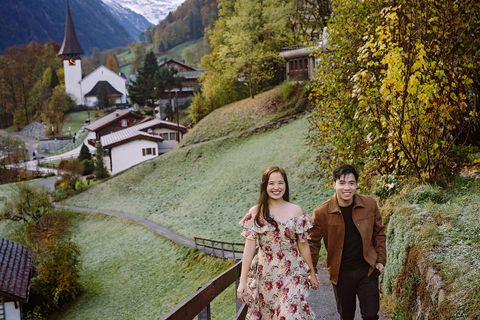 Couple photoshoot in Lauterbrunnen, Switzerland: couple holding hands and walking on a hillside path with the valley church, autumn trees, and snowy Alps behind.