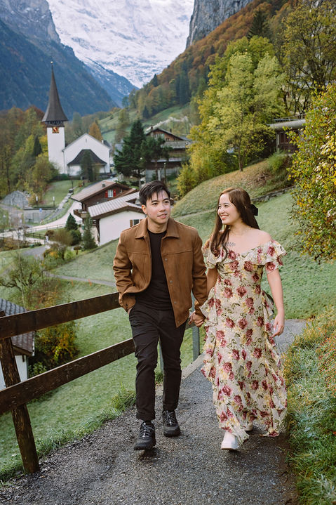 Couple photoshoot in Lauterbrunnen, Switzerland: couple holding hands and walking on a hillside path with the valley church, autumn trees, and snowy Alps behind.