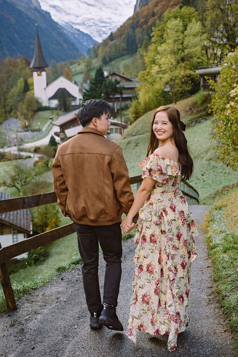 Couple photoshoot in Lauterbrunnen, Switzerland: couple holding hands and walking on a hillside path with the valley church, autumn trees, and snowy Alps behind.