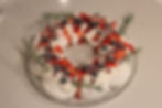 A festive pavlova wreath with whipped cream, berries, and red sauce, garnished with rosemary on a glass platter, set on a marble surface.