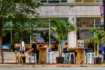 People dining at rustic wooden tables and bright blue stools on a widened downtown sidewalk, surrounded by potted palm trees and historic storefronts.