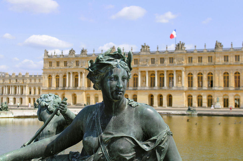 Bronze statue of a woman with flowing hair and draped clothing in front of the Palace of Versailles, with the French flag visible atop the building and a reflecting pool in the background.