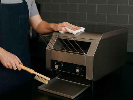 Barista cleaning a commercial conveyor toaster in a café kitchen