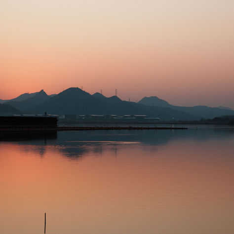 A calm lake reflecting a golden sunset with silhouetted mountains in the distance, still water and warm light, no people