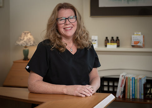 A woman with red curly hair and glasses leans on the side of a desk and smiles.