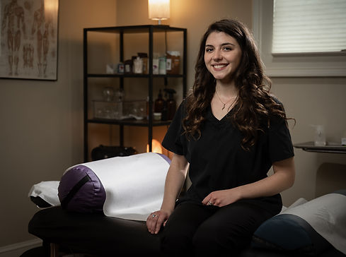 A woman with long brown hair smiles and sits at the edge of a massage table.