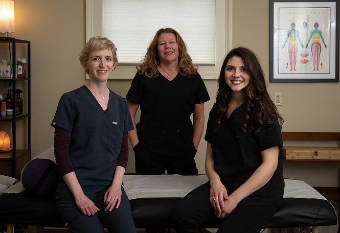 Three women in a treatment room. The women on the ends sit on a massage table, and the woman in the middle stands. A window covered by a blind is behind them.