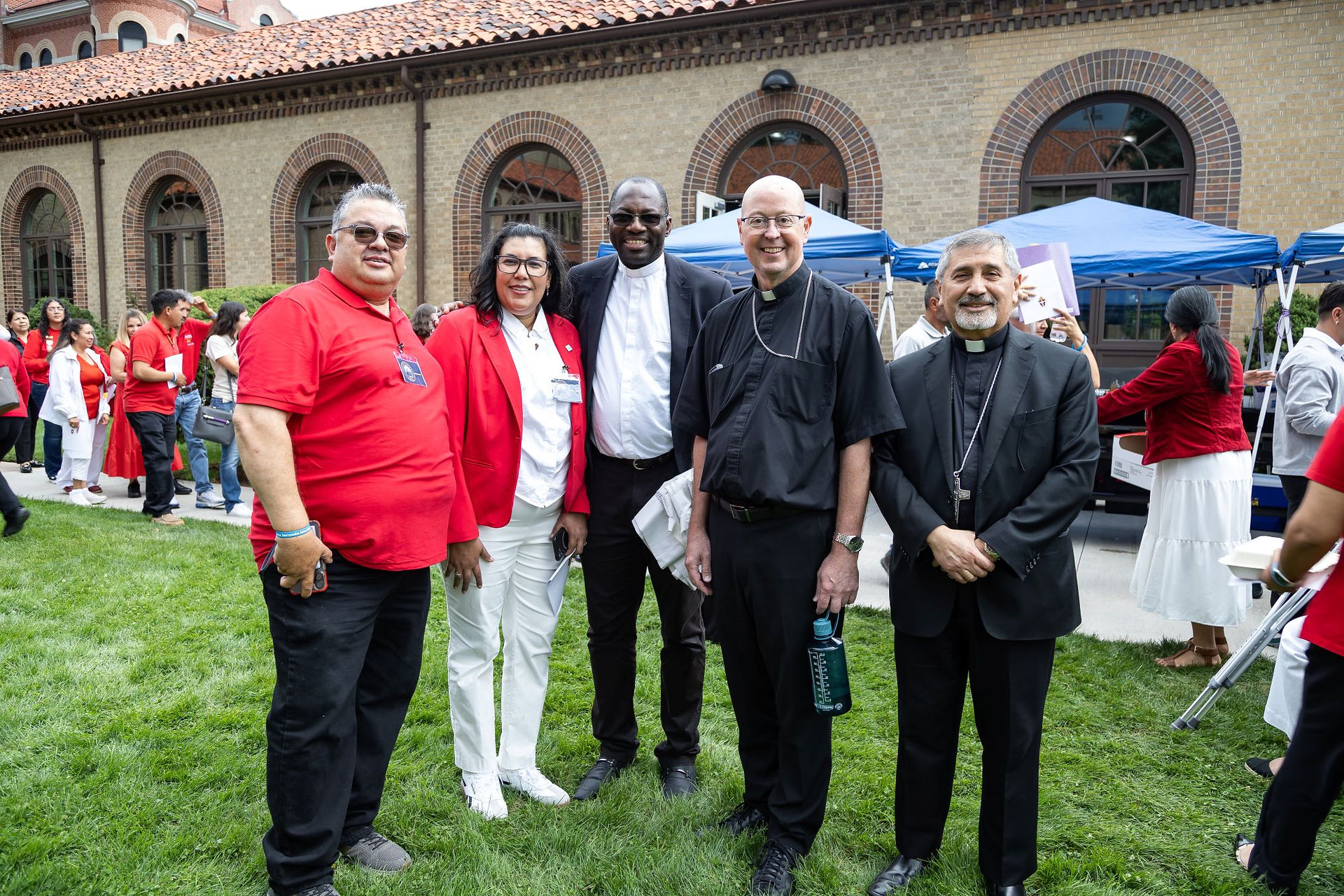 Five smiling attendees, including clergy, pose on a lawn outside a brick church with blue tents at a cheerful gathering.