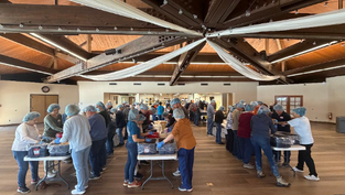 Personas con gorras azules empacan alimentos en mesas, dentro de un salón con techo de vigas oscuras. Ambiente colaborativo y organizado.