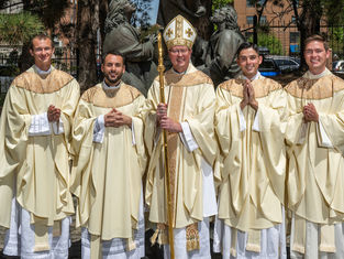 Cinco clérigos católicos con vestiduras crema posan al aire libre; el obispo central sostiene un báculo frente a una estatua.