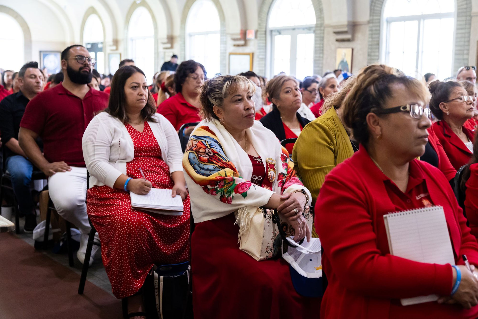 Crowd of adults in red and white sits attentively in a bright arched hall, some holding notebooks, during a formal gathering.