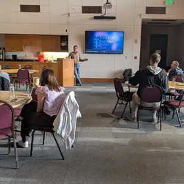 Personas escuchando a un orador en una sala con sillas moradas. En la pantalla: "VIRTUS: Excellence Builds Trust". Ambiente tranquilo.