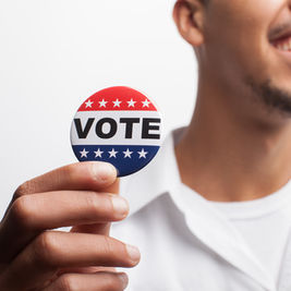 Hombre sonriente sostiene un botón rojo, blanco y azul que dice VOTE, sobre fondo blanco.