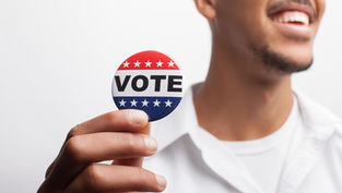 Hombre sonriente sostiene un botón rojo, blanco y azul que dice VOTE, sobre fondo blanco.