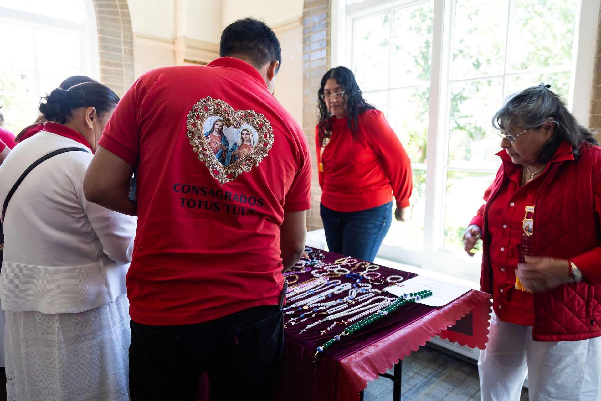 Group in red shirts examines rosaries on a table in a bright room; one shirt reads CONSAGRADOS TOTUS TUUS.