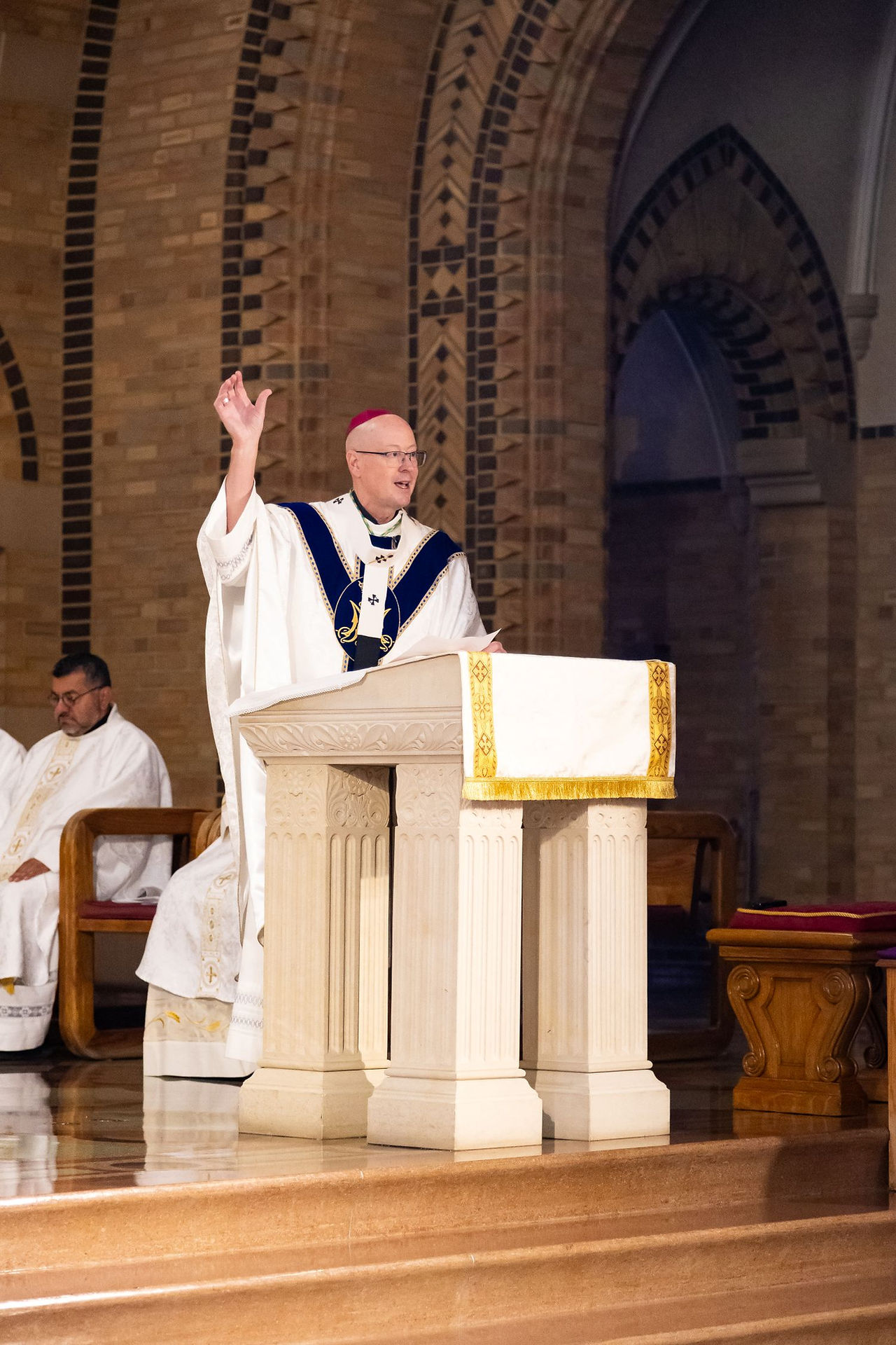 Priest in white and blue vestments raises a hand while speaking at a church lectern, with another clergyman seated behind.