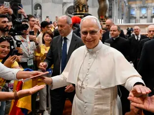 Un clérigo sonriente en túnica blanca saluda a una multitud en una iglesia grande. Personas toman fotos; ambiente alegre y colorido.