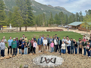 Grupo grande de familias y niños posando sonrientes en un campamento montañoso con pinos, cabañas y montañas al fondo.