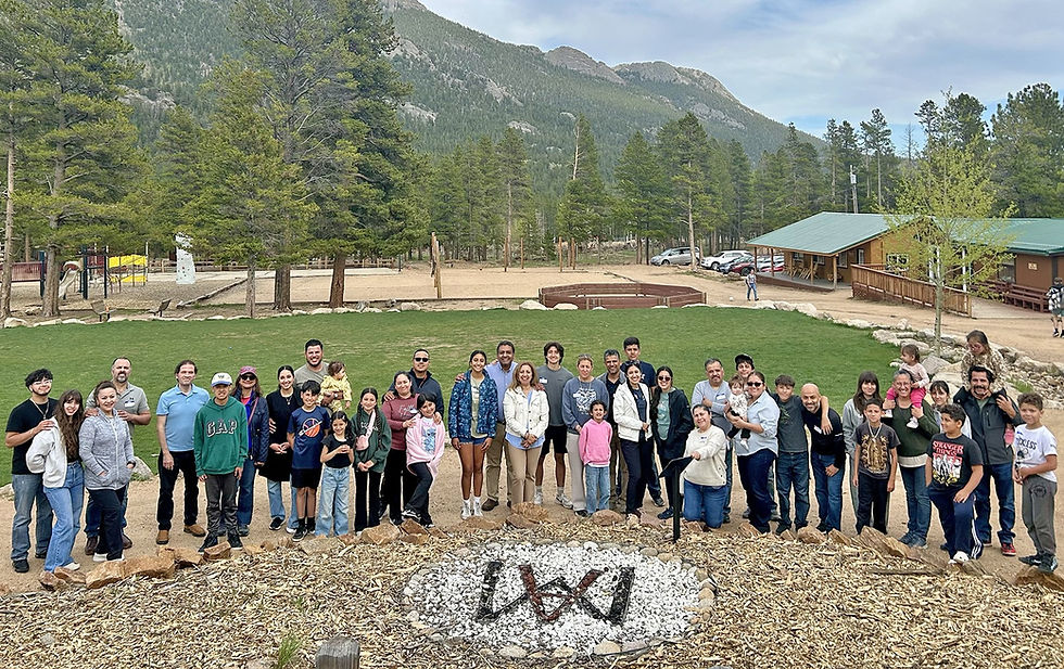 Grupo grande de familias y niños posando sonrientes en un campamento montañoso con pinos, cabañas y montañas al fondo.