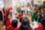 Church congregation claps and sings around a guitarist, with priests in green vestments in a bright hall.