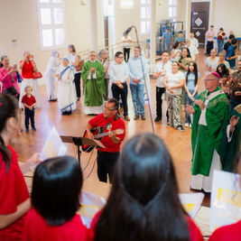 Church congregation claps and sings around a guitarist, with priests in green vestments in a bright hall.