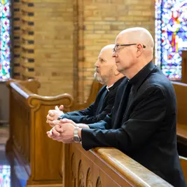 Dos hombres con vestimenta religiosa rezan en un banco de iglesia con vitrales coloridos en el fondo. Ambiente solemne.