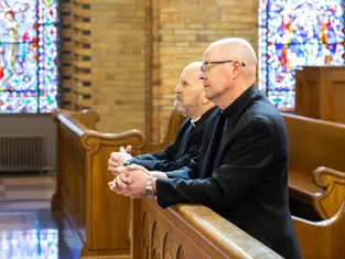 Dos hombres con vestimenta religiosa rezan en un banco de iglesia con vitrales coloridos en el fondo. Ambiente solemne.