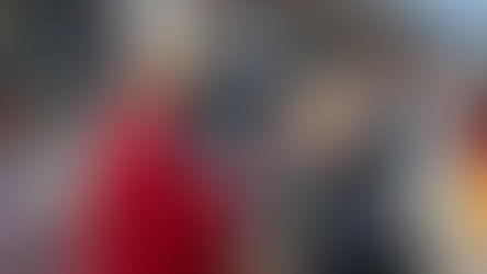 Un obispo con túnica roja y mitra bendice a una mujer que sostiene hojas. Fondo urbano con transeúntes desenfocados. Ambiente solemne.