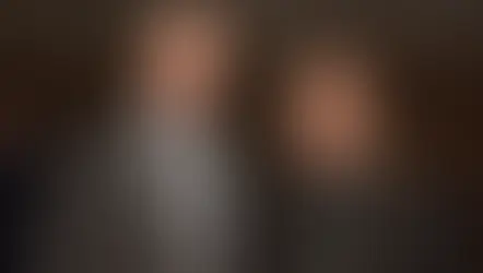 Dos hombres sonrientes en trajes formales en una sala de conferencias. Fondo oscuro con personas borrosas. Uno lleva un collar religioso.