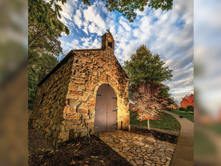 Pequeña capilla de piedra con puerta de madera, rodeada de árboles en un entorno otoñal. Cielo azul con nubes dispersas. Ambiente tranquilo.