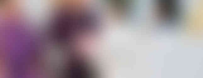 A bishop in purple vestments sits, holding a book, with a man in white kneeling. Flowers and a decorative chair are in the church setting.